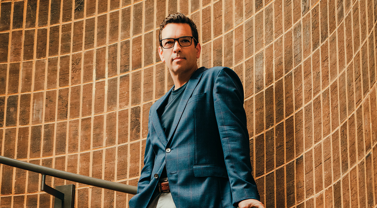 Dressed in a dark blue T-shirt, a blue blazer, and beige slacks, Robert Ford leans casually against the handrail on a curving staircase.